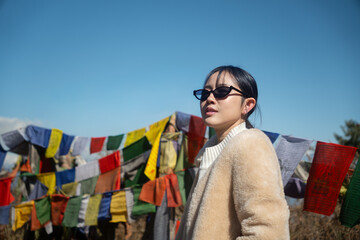 A young Asian woman tourist wearing sunglasses and a warm cream fleece coat looking away, standing in front of traditional colorful Buddhist prayer flags on a mountain under a clear blue sky © Oil And Den