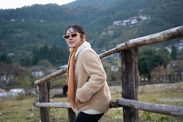 A young Asian woman tourist wearing sunglasses, a warm cream fleece coat, and an orange scarf smiling at the camera while leaning against a rustic wooden fence in a countryside valley with a mountain  © Oil And Den