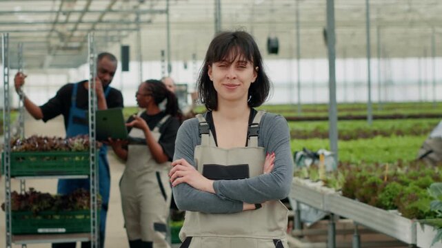 Confident professional portrait of woman posing with arms crossed in greenhouse while farm workers are using, showing professional greenhouse work and organic farming in a controlled greenhouse.