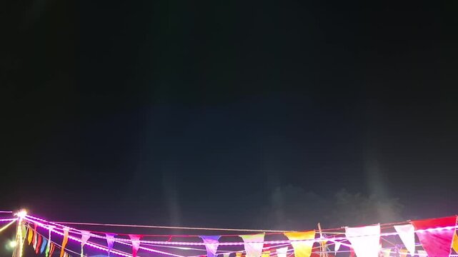 A wide shot of layered, multi-colored triangular festival flags illuminated by bright string lights at a night market fair, featuring subtle searchlight beams shooting up into the misty dark sky.