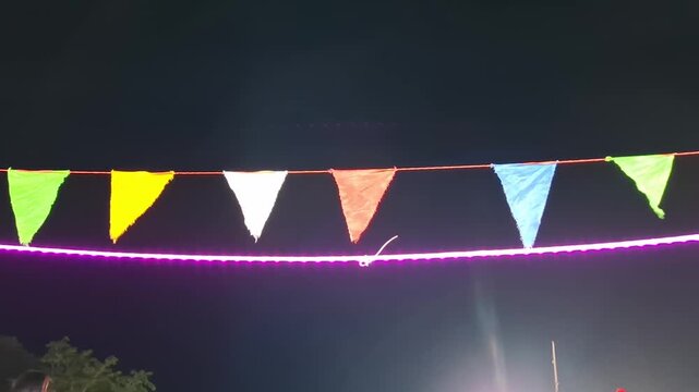 A close-up shot of vibrant, multi-colored triangular festival bunting flags hanging on a string line with a bright pink neon LED light tube glowing underneath against a dark night sky background