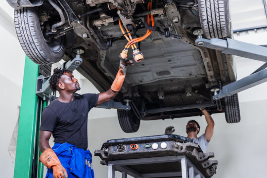 Professional EV technicians inspecting high-voltage electrical connectors beneath a lifted electric vehicle during battery pack removal and maintenance.