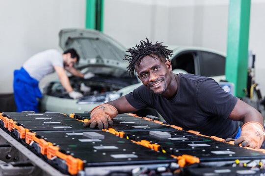 Confident electric vehicle technician smiling beside a high-voltage battery pack during maintenance in a modern EV service center.