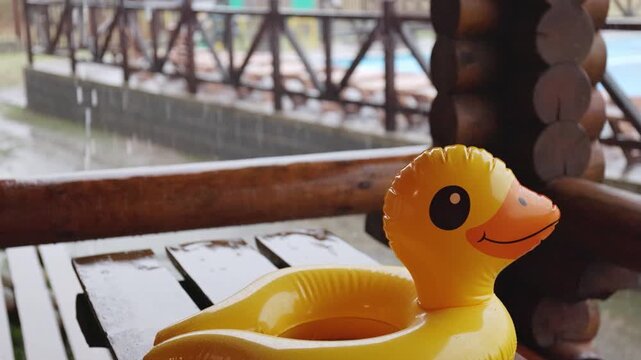 An inflatable duck lies on wooden table in gazebo at recreation center against the backdrop of pool covered with rain