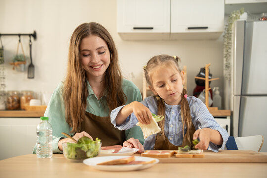 Cute girl using both hands to build a sandwich while her smiling mother helps in the kitchen. Happy mother and daughter preparing healthy breakfast sandwiches together on a wooden cutting board.