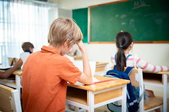 Schoolboy sitting at his desk, holding his forehead while concentrating or thinking in class. Elementary student studying at his classroom desk with classmates and a green chalkboard in background.