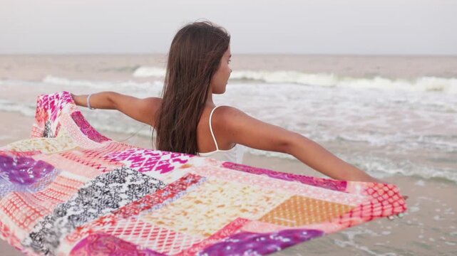 A girl in a summer T-shirt and shorts is walking along the beach looking at the horizon and holding a shawl in her hands