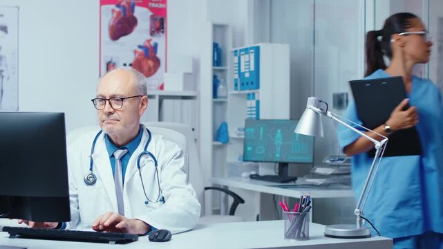 Doctor looks toward the camera after typing at a computer in a glass walled hospital clinic while a nurse speaks with a patient behind him.
