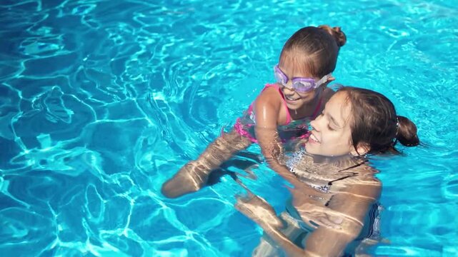 Two girls in bright bikinis are playing together in a pool with blue water on a summer day on a long-awaited vacation