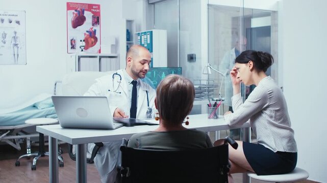Daughter talks with a doctor about recovery for her elderly mother in a wheelchair, focusing on treatment planning and mobility support.