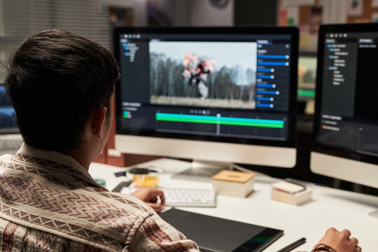 Over the shoulder shot of man editing visual effects footage on desktop monitors, managing post production workflow for digital content creation. Useful for media agency, video editing, creative tech