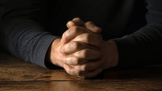 Close up of interlocked hands in a prayerful gesture resting on a dark wooden table surface under dramatic low key lighting for spiritual reflection