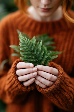 Close up of a woman in orange knitted sweater gently holding green fern leaves in her hands.