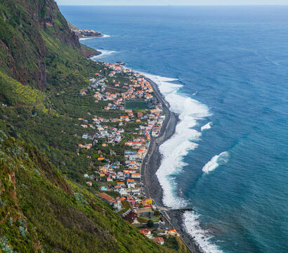 A high-angle shot capturing the coastal village of Paul do Mar nestled tightly between towering green cliffs and the ocean, Madeira, Portugal.