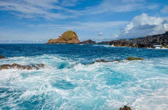Powerful white ocean waves foaming and crashing into dark volcanic rock reefs near Porto Moniz natural pools, Madeira, Portugal.