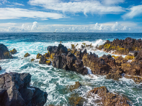 Powerful white ocean waves foaming and crashing into dark volcanic rock reefs near Porto Moniz natural pools, Madeira, Portugal.