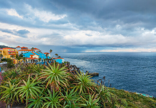 Colorful residential houses on a coastal hill looking over a dramatic dark stormy ocean under a cloudy sky in Canico, Madeira, Portugal.