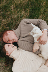 Fototapeta premium A joyful couple lies on grass, cuddling a goat kid, sharing a tender outdoor moment, surrounded by warm sunlight and soft sweaters, capturing simple, everyday happiness and connection.