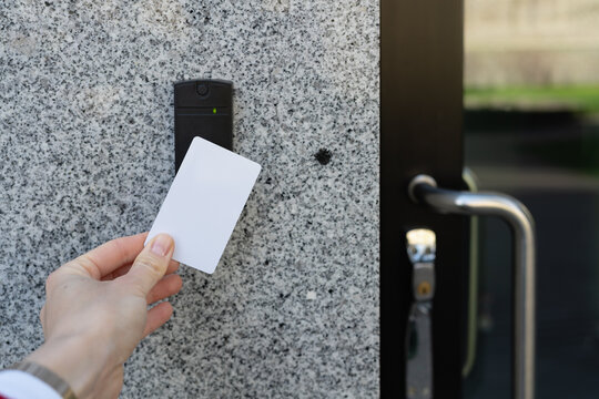 Closeup of a person's employee's hand holding a white access key pass card near a reader access device on a grey granite wall and a black glass door locked door, green light indicates granted access, 