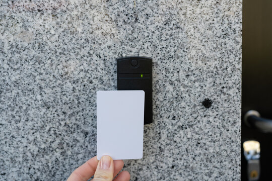 Closeup of a person's employee's hand holding a white access key pass card near a reader access device on a grey granite wall and a black glass door locked door, green light indicates granted access, 