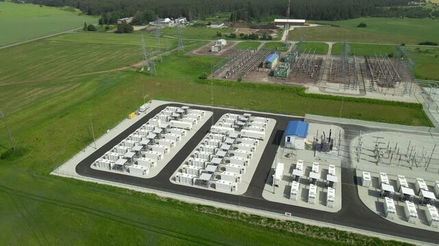 High angle drone footage showing a modern BESS facility featuring rows of industrial containers and a high voltage electrical substation, showcasing clean energy infrastructure in a green field