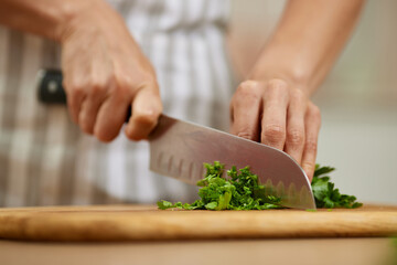 Fototapeta premium Chef's hands holding a knife, finely chopping fresh green parsley herbs on a rustic wooden cutting board in a kitchen