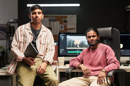 Portrait of two young men posing in video editing studio, holding laptop and sitting by desktop monitor. Useful for creative agency, content production, teamwork marketing