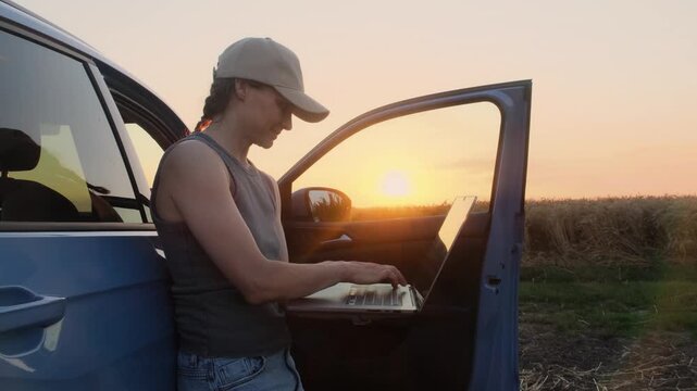 A young farmer woman in work clothes stands next to the car in the fields at sunset and enters information about the harvest into her laptop. She is tired, but satisfied with the result of her work.