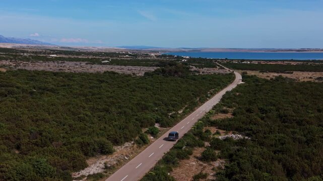 Silver camper van car driving down a long road through green bushes on Pag Island, Croatia. Aerial.