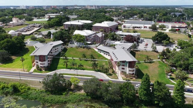 Florida Southern College campus showing Frank Lloyd Wright architecture in Lakeland, Florida, with covered walkways linking buildings amid palm trees near lakeside road. Aerial drone orbit shot