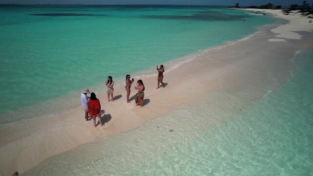 High-angle drone shot of stylish group enjoying a luxury beach experience, taking photos and laughing on a remote white sandbank in Cayo de Agua Los Roques