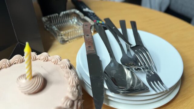 Close-up of a frosted chocolate birthday cake with an unlit candle beside a stack of white plates and metal silverware. The party dessert setting rests on a wooden table in a bright indoor home.