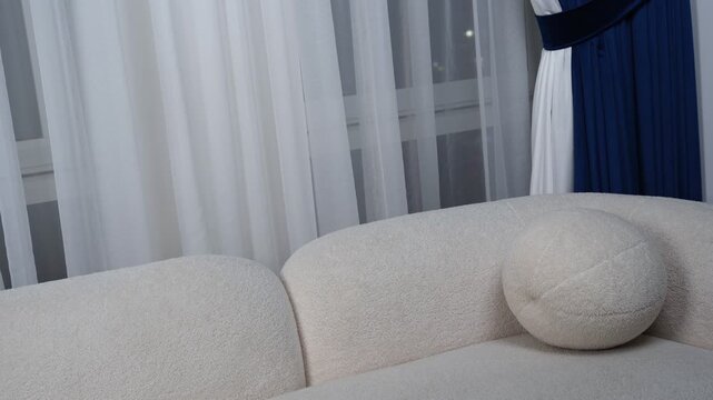 Static shot of a modern white textured boucle sofa featuring a round spherical pillow. The elegant indoor furniture sits in front of white sheer window curtains and dark blue drapes at night.