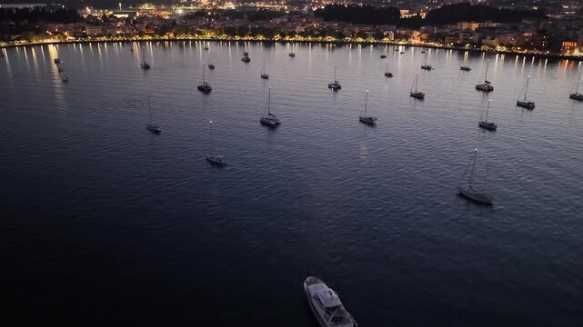 Aerial footage capturing the twilight atmosphere in Corfu bay. Multiple yachts and sailboats rest on the calm water as the city lights begin to illuminate the coastline, creating golden reflections on