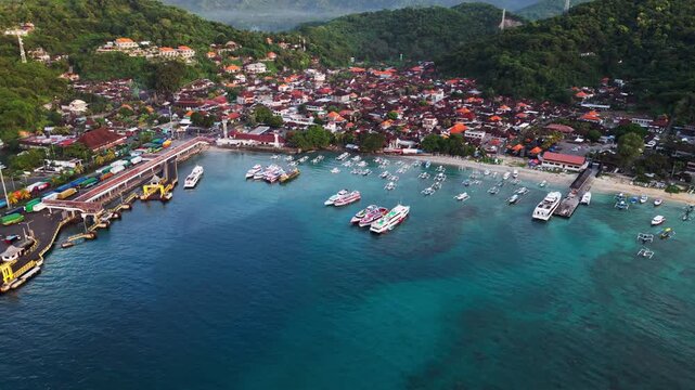Padang Bai coastal harbor in Bali featuring anchored traditional fishing boats and speedboats floating on vibrant turquoise ocean water, pullback aerial drone footage