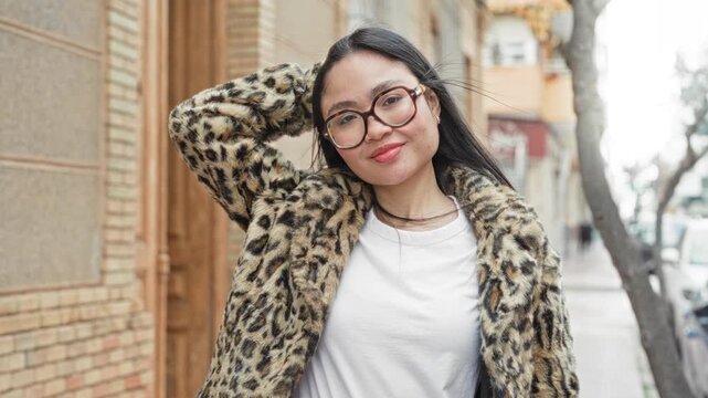 Woman wearing glasses and a leopard fur coat smiles with hand behind head on street, leaning against a building; casual confidence.