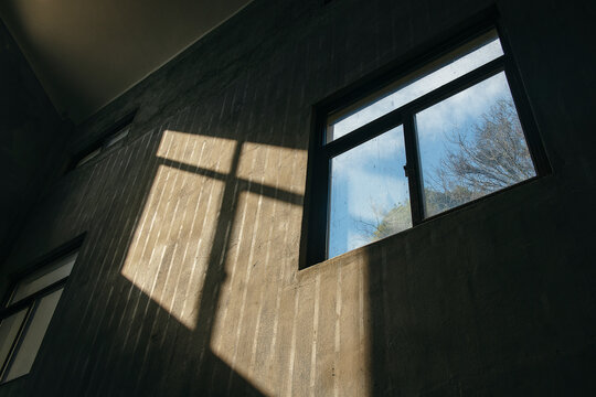Sunlight casting a cross-shaped shadow through a window onto a textured concrete wall with a view of the blue sky outside