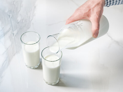Man's hand pours fresh milk from a glass bottle into a clear glass on a white marble table. Symbolizes dairy nutrition, healthy breakfast, and organic farming. Ideal for food and wellness content