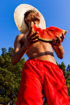 Young man enjoying summer feeling, eating watermelon outdoors under blue sky