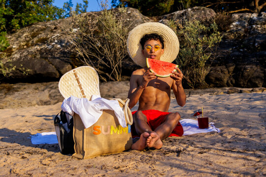 Young man enjoying summer vacation on sandy beach, eating watermelon