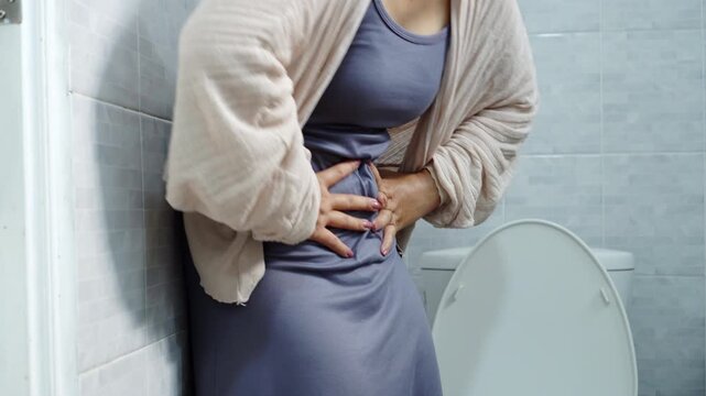 woman suffering from severe stomach ache, food poisoning, or diarrhea, holding her painful belly in front of a toilet bowl in the bathroom. Healthcare and medical concept