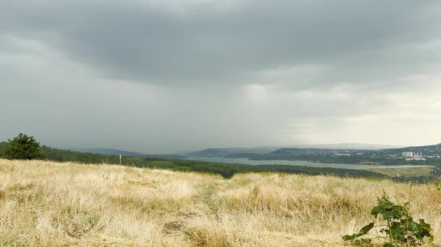 A refreshing summer shower pours down over the quiet reservoir and green hills of Simferopol, Crimea. The dramatic overcast sky creates a gloomy yet deeply peaceful atmosphere.