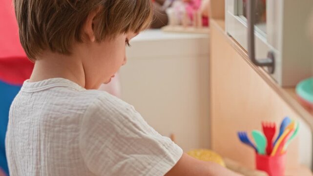 Boy holds a toy burger and colorful play utensils at a play kitchen inside a building; playful curiosity.