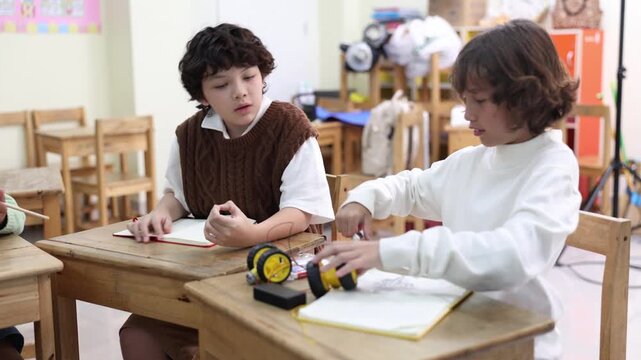 Two elementary school boys sit at wooden desks in a bright classroom. One smiling student plays with a small wheeled toy project while his classmate watches curiously during a lesson.