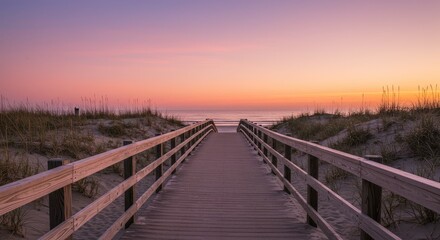 Obraz premium A serene wooden boardwalk leads through beach grass to the calm ocean at sunset.