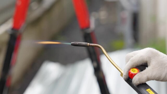 Close-up of a worker holding a gas blowtorch with an open orange flame outdoor. Industrial roofing or construction process, plumbing heating torch tool concept