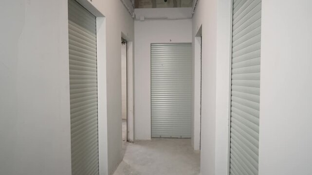 Symmetrical hallway with white walls gray roller doors numbered above each unit. Neutral color palette with soft overhead lighting. Clean industrial aesthetic suitable for logistics commercial use