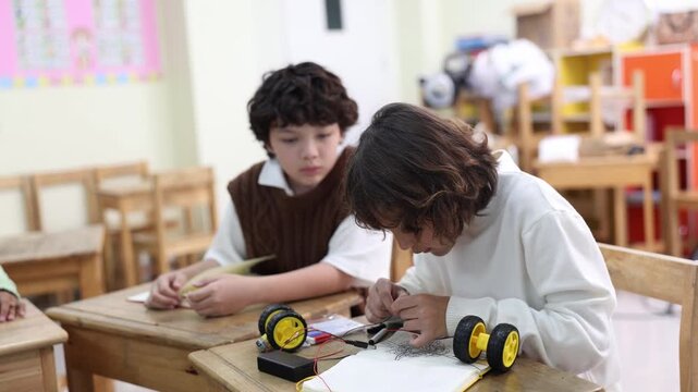 Two elementary school boys sit at wooden desks in a bright classroom. One smiling student plays with a small wheeled toy project while his classmate watches curiously during a lesson.
