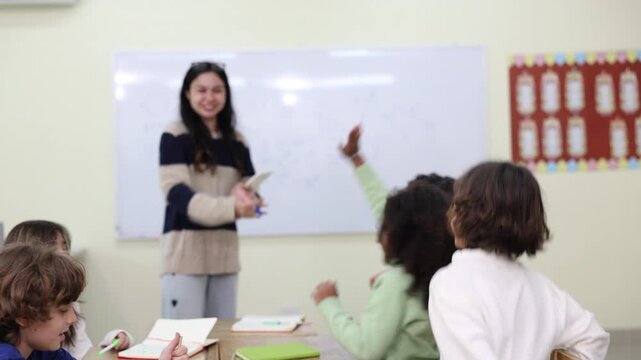A young female teacher holds a digital tablet and marker while enthusiastically explaining a math lesson to a diverse group of elementary students in a modern classroom.