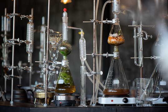 Close-up of complex glass laboratory distillation setups with flasks containing green herbs and brown liquid heated on hot plates.